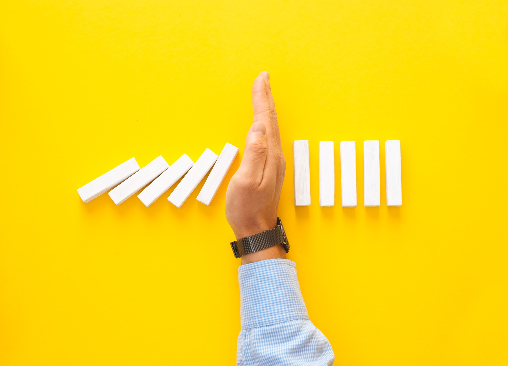 Businessman hand stopping falling dominos on yellow background. Business management crisis concept.